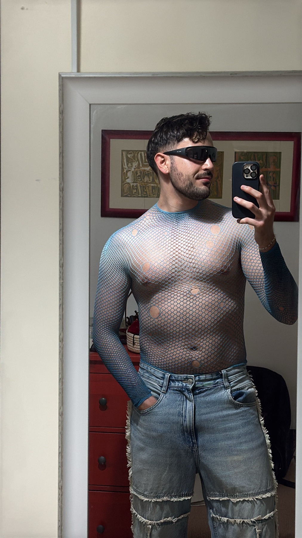 A man in a blue mesh top and distressed jeans takes a mirror selfie while wearing sunglasses.