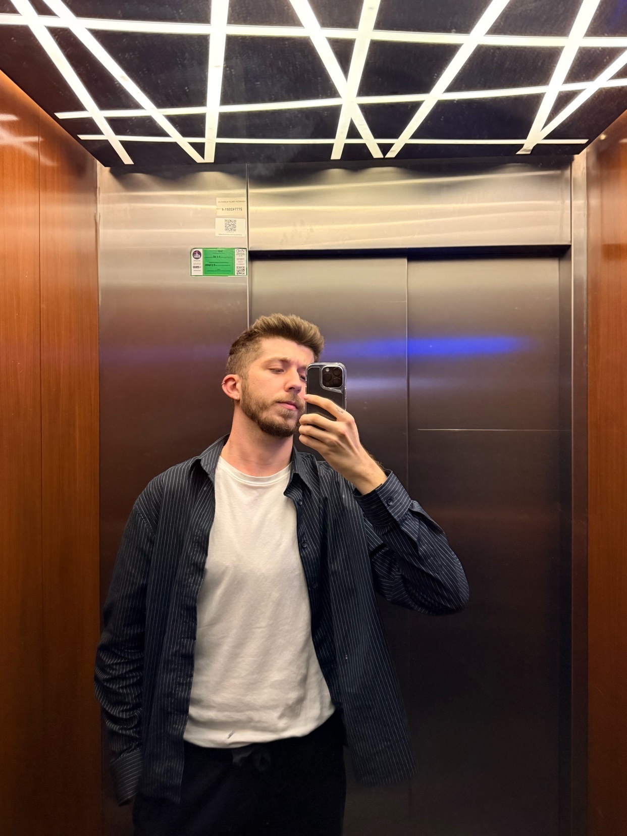 A man takes a selfie in an elevator. He is shirtless and wearing an open button-down shirt.