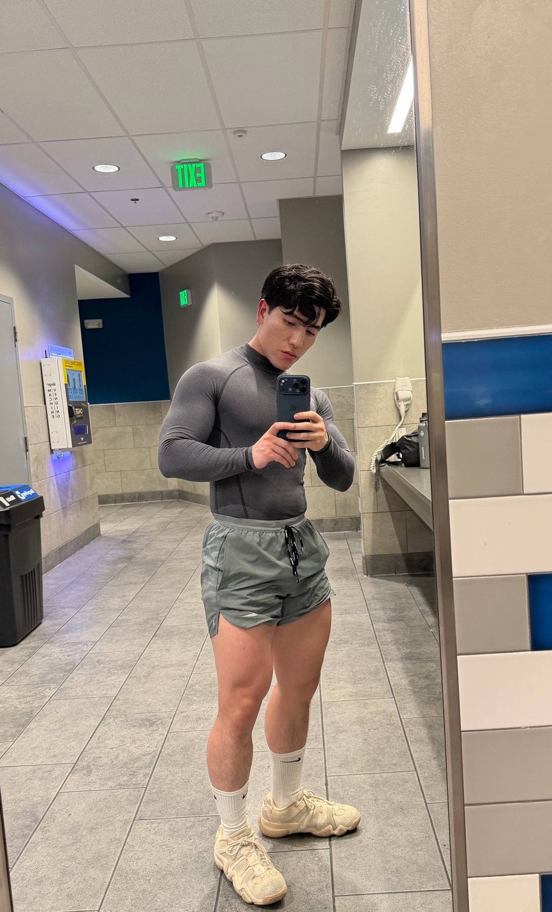 A man takes a mirror selfie at the gym. He is wearing a long-sleeved shirt and shorts, showcasing his muscular physique.