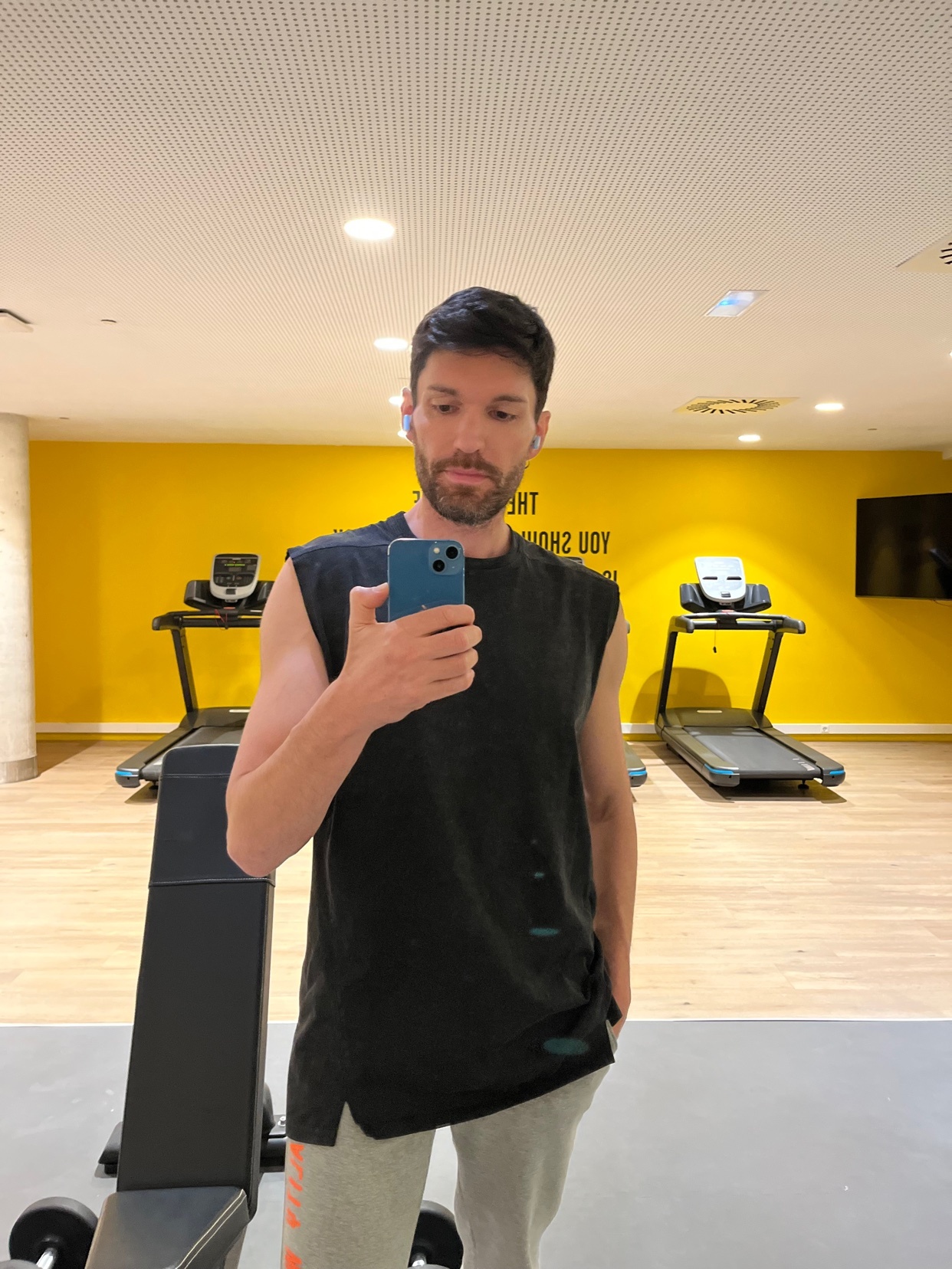 A man takes a selfie in a gym, wearing a sleeveless shirt and sweatpants. Treadmills are visible in the background.