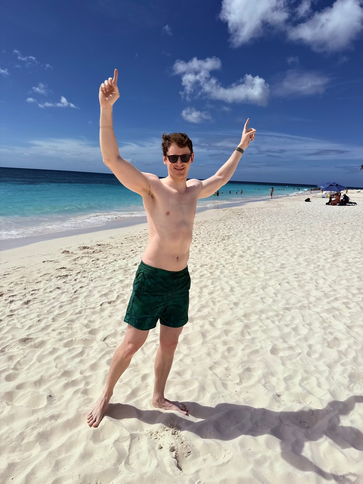 A man stands on a white sand beach with his arms raised. The clear blue ocean and sky create a beautiful backdrop.