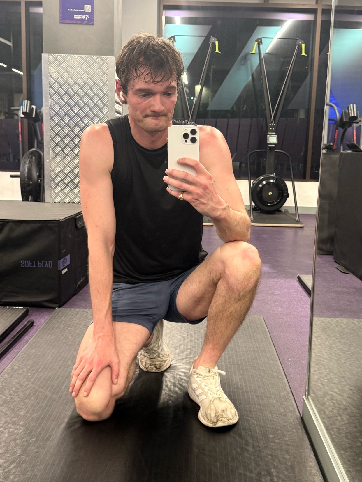 A man takes a mirror selfie at the gym. He is shirtless and wearing shorts and sneakers.