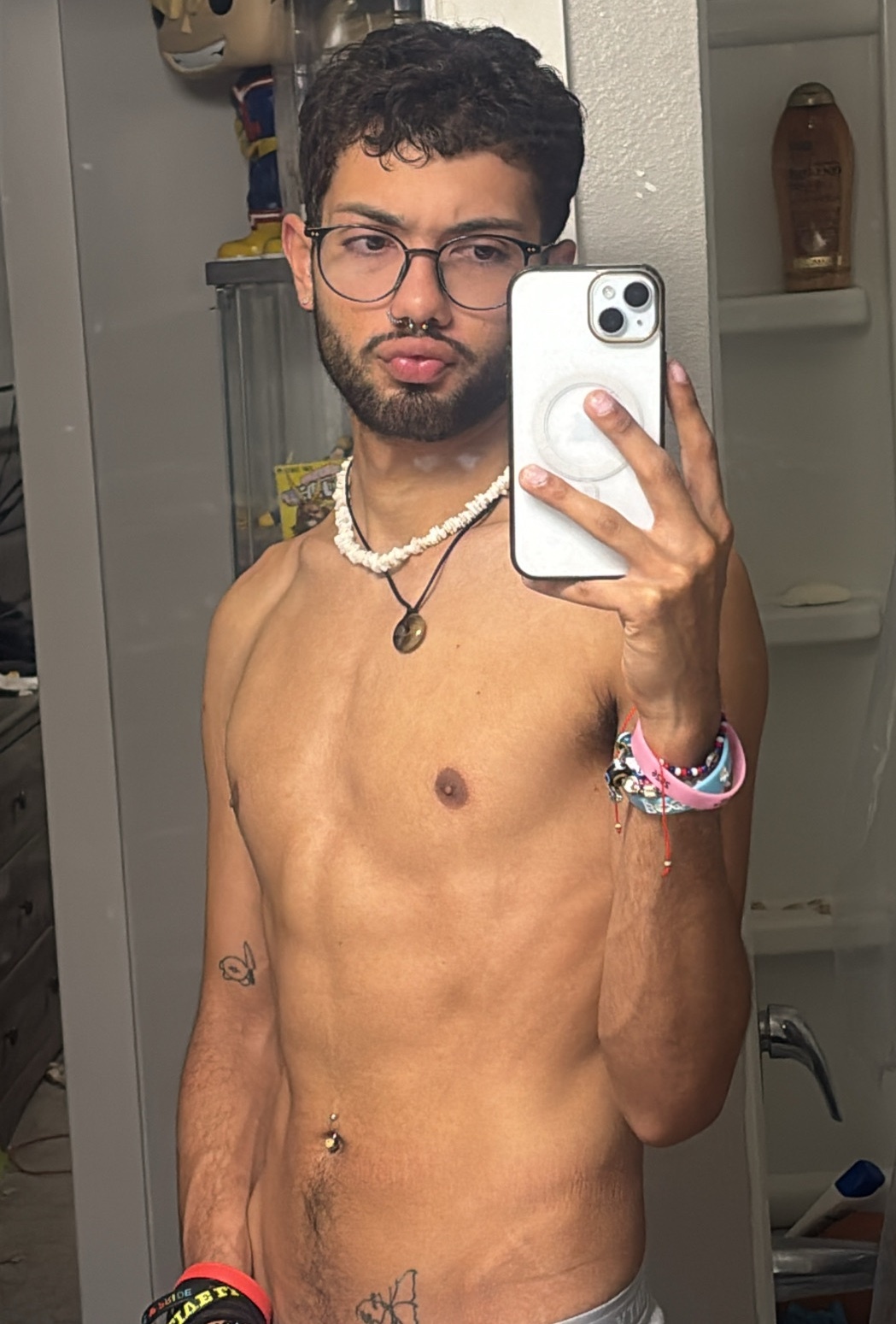A young man with curly hair and glasses takes a selfie in a bathroom. He is wearing a necklace and bracelets, and has tattoos on his arm and chest.