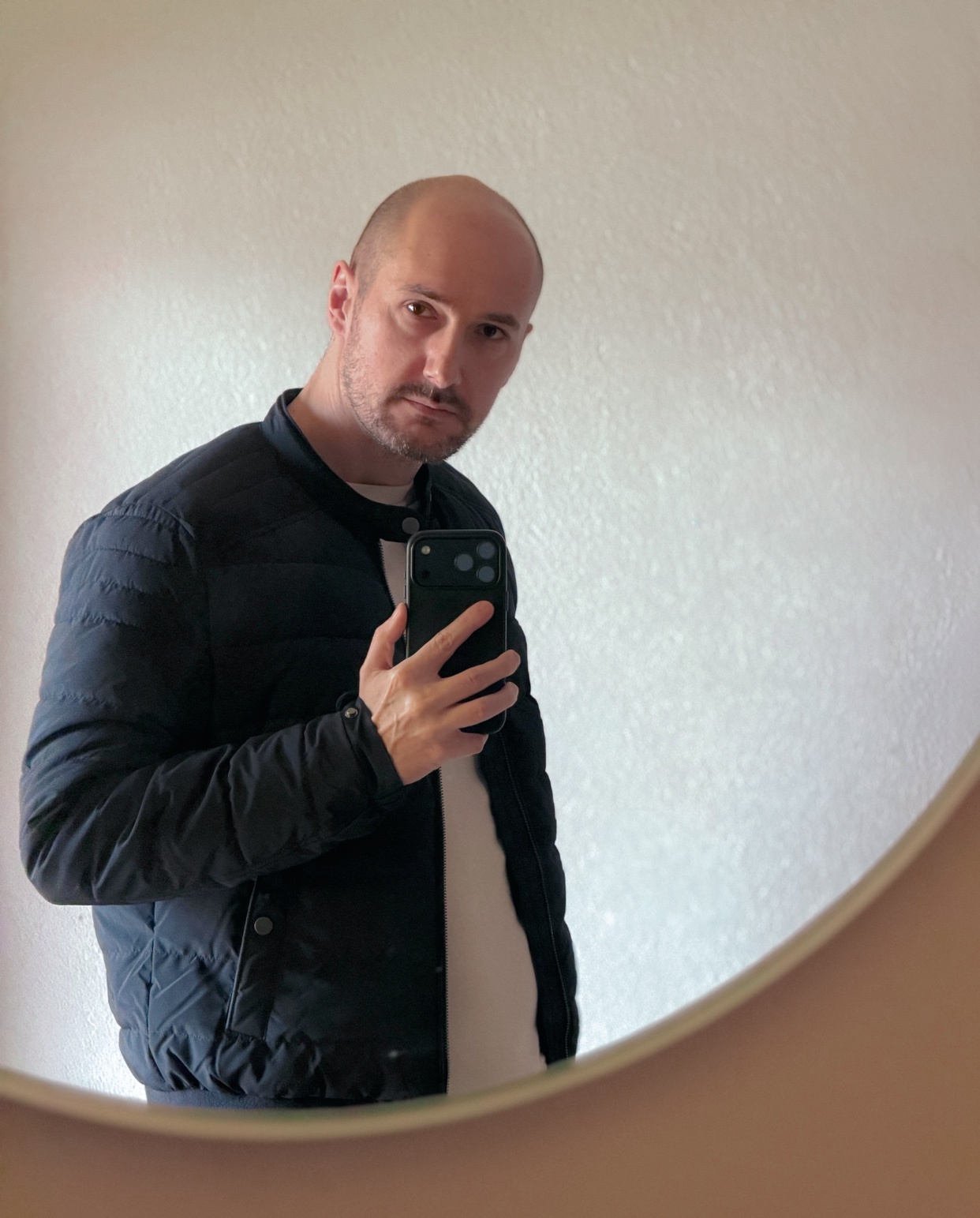 A man with a bald head and stubble takes a mirror selfie while wearing a black puffy jacket over a white t-shirt.