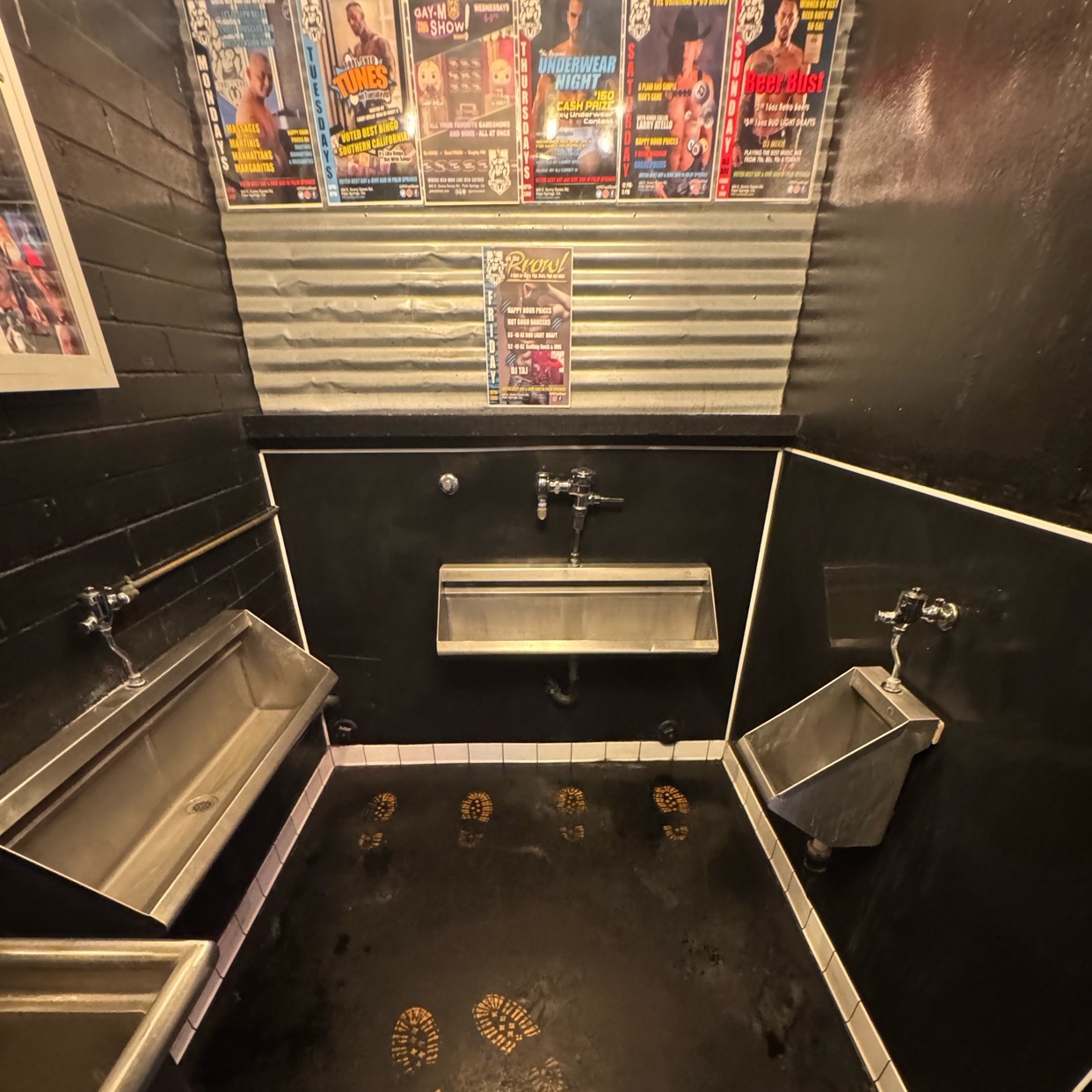 A grungy, industrial-style public restroom with multiple urinals and posters on the wall. Footprints are visible on the dark, wet floor.
