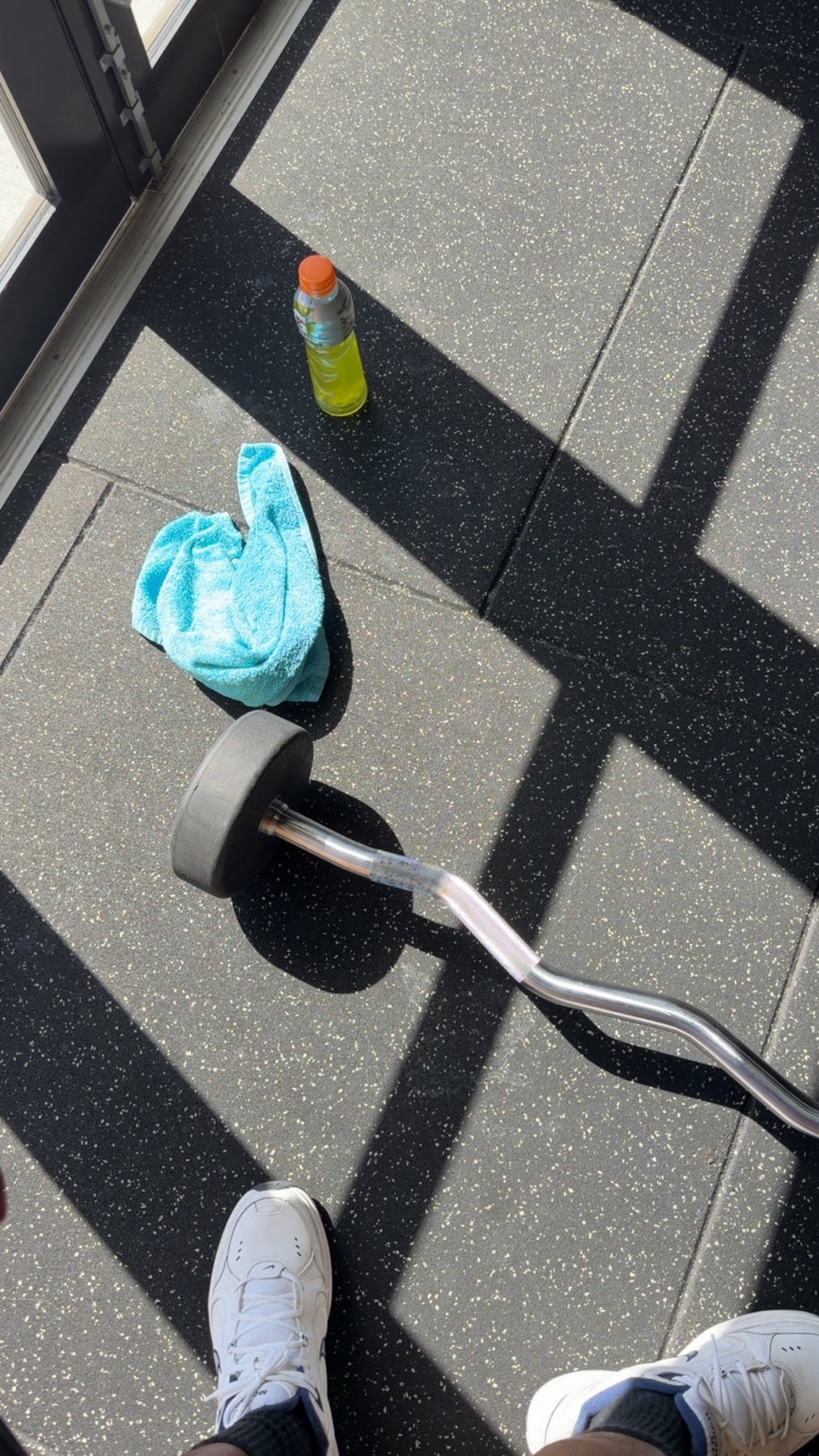 A barbell, a light blue towel, and a bottle of sports drink sit on a dark gym floor, with strong shadows cast across the scene.