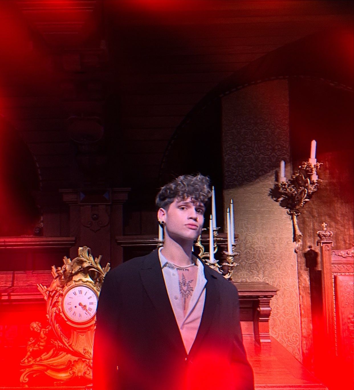 A man poses in a dark room with a vintage clock and candles. He is wearing a suit and is shirtless.