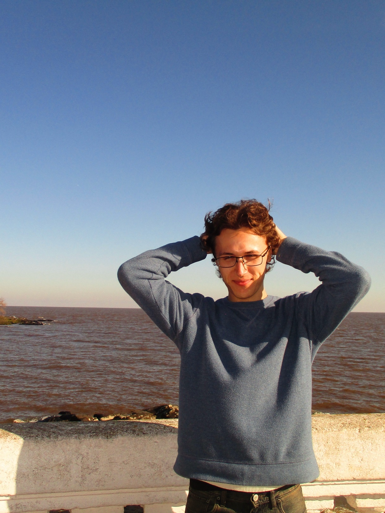 A man with curly hair and glasses poses with his hands behind his head, wearing a blue sweater and jeans. He stands outdoors with a clear blue sky and a body of water behind him.