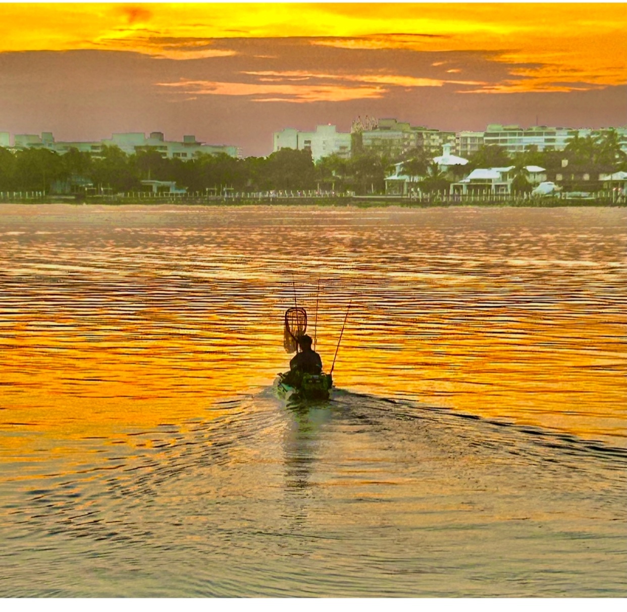 A lone figure in a kayak fishes on a body of water during a vibrant sunset. The golden light reflects off the water, with a cityscape visible in the distance.