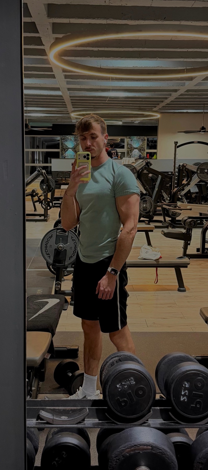 A man takes a mirror selfie at the gym, showcasing his physique. He is wearing a t-shirt and shorts, with weights visible in the foreground.