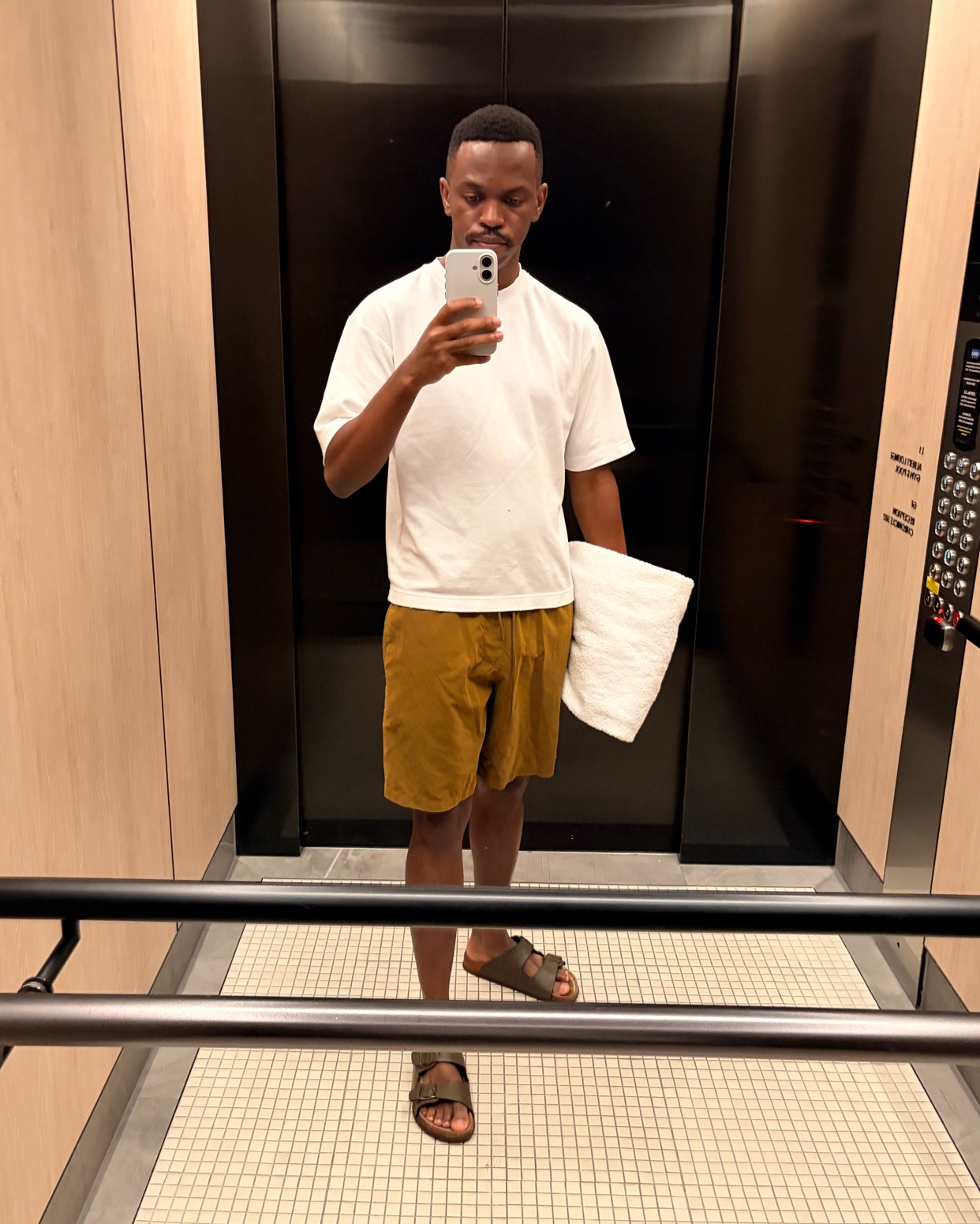 A man takes a selfie in an elevator while holding a towel. He is wearing a white t-shirt, brown shorts, and sandals.