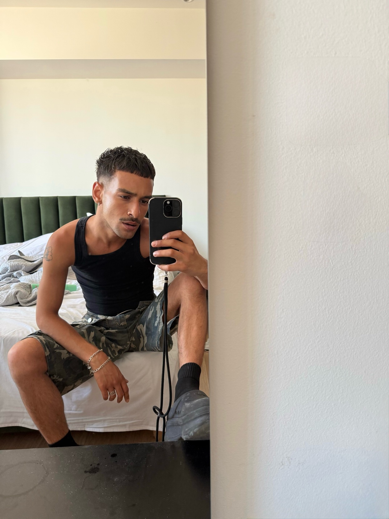 A man takes a mirror selfie while sitting on a bed. He is shirtless and wearing camouflage shorts.