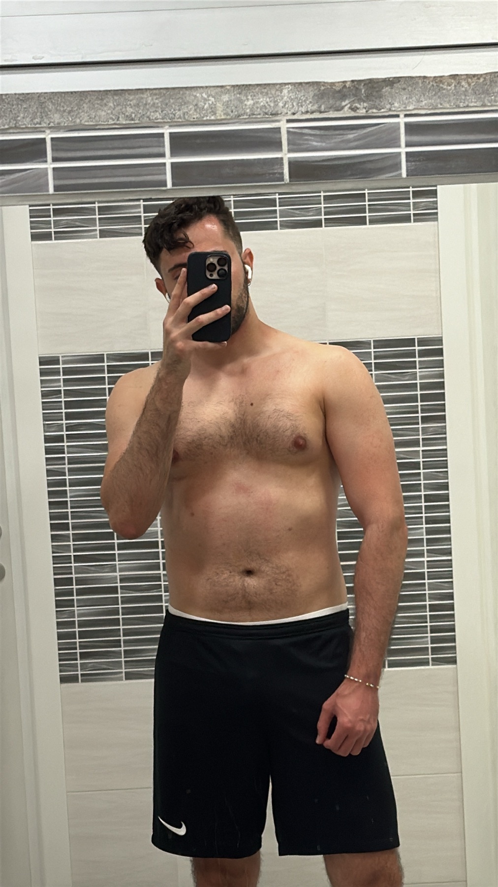 A man takes a mirror selfie in a bathroom, wearing black Nike shorts and no shirt. He has short dark hair, a beard, and is wearing earbuds and an arm bracelet.