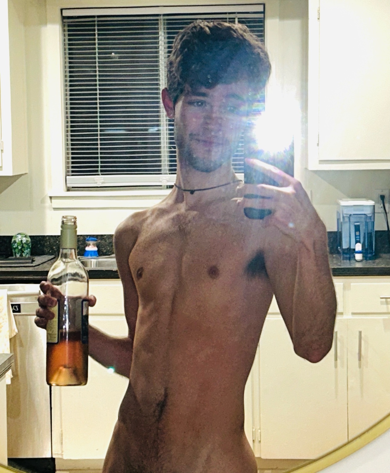 A man takes a mirror selfie, shirtless, holding a bottle of wine in a kitchen. The image highlights his physique and the casual setting.