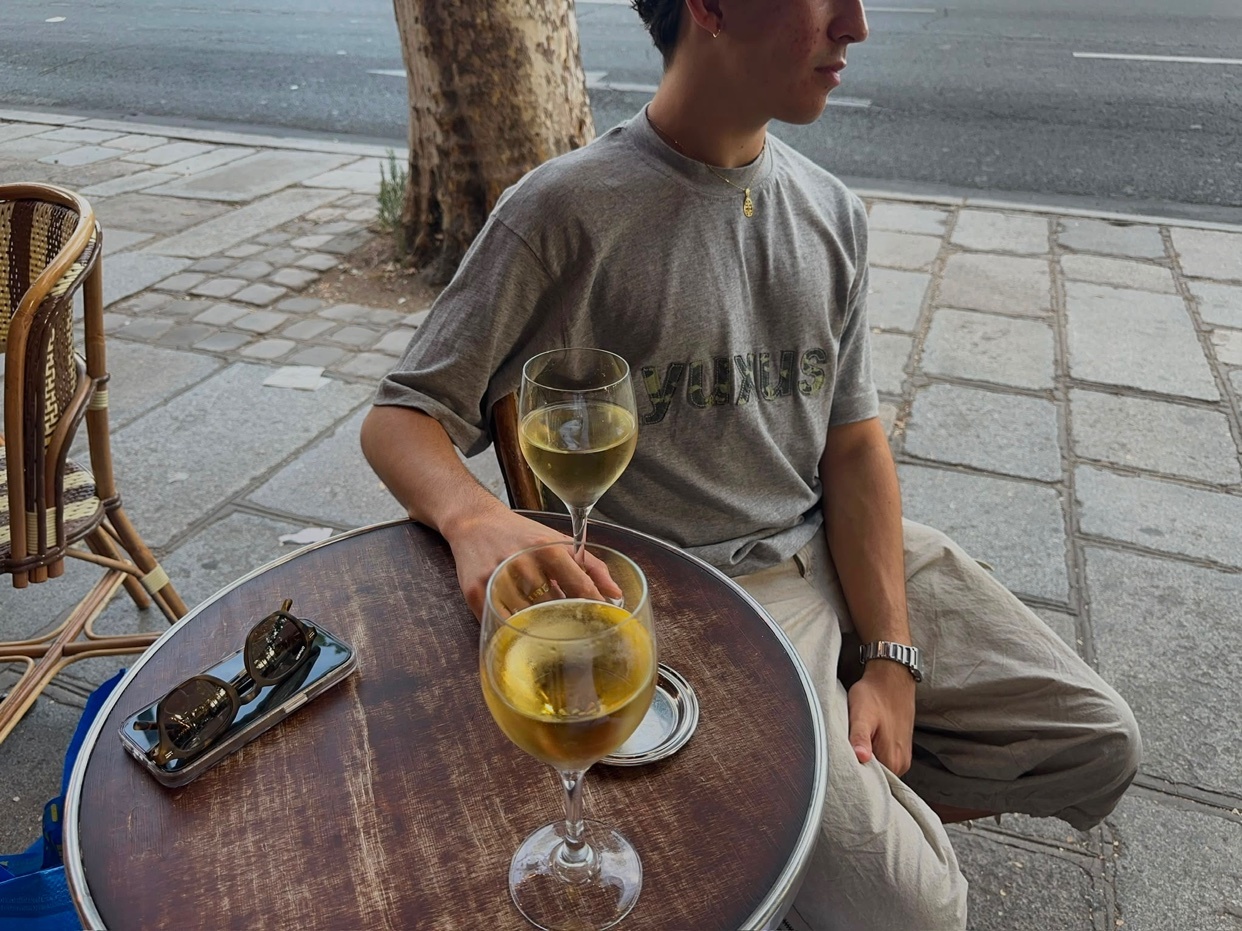 A man sits at a table outside a cafe with two glasses of white wine and a pair of sunglasses on the table.