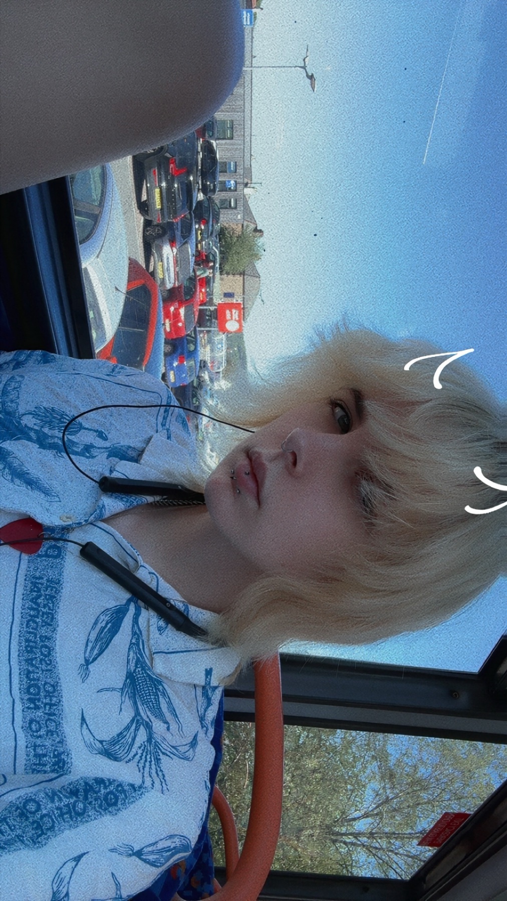 A young woman with blonde hair and blue eyes takes a selfie on a bus. She is wearing a blue and white button-up shirt and has earbuds around her neck.