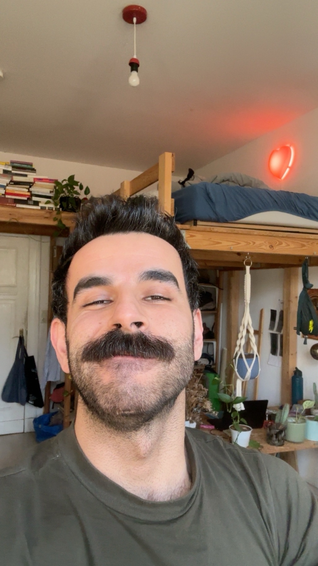 A man with a large mustache smiles at the camera. The photo is taken in a bedroom with a loft bed and plants.