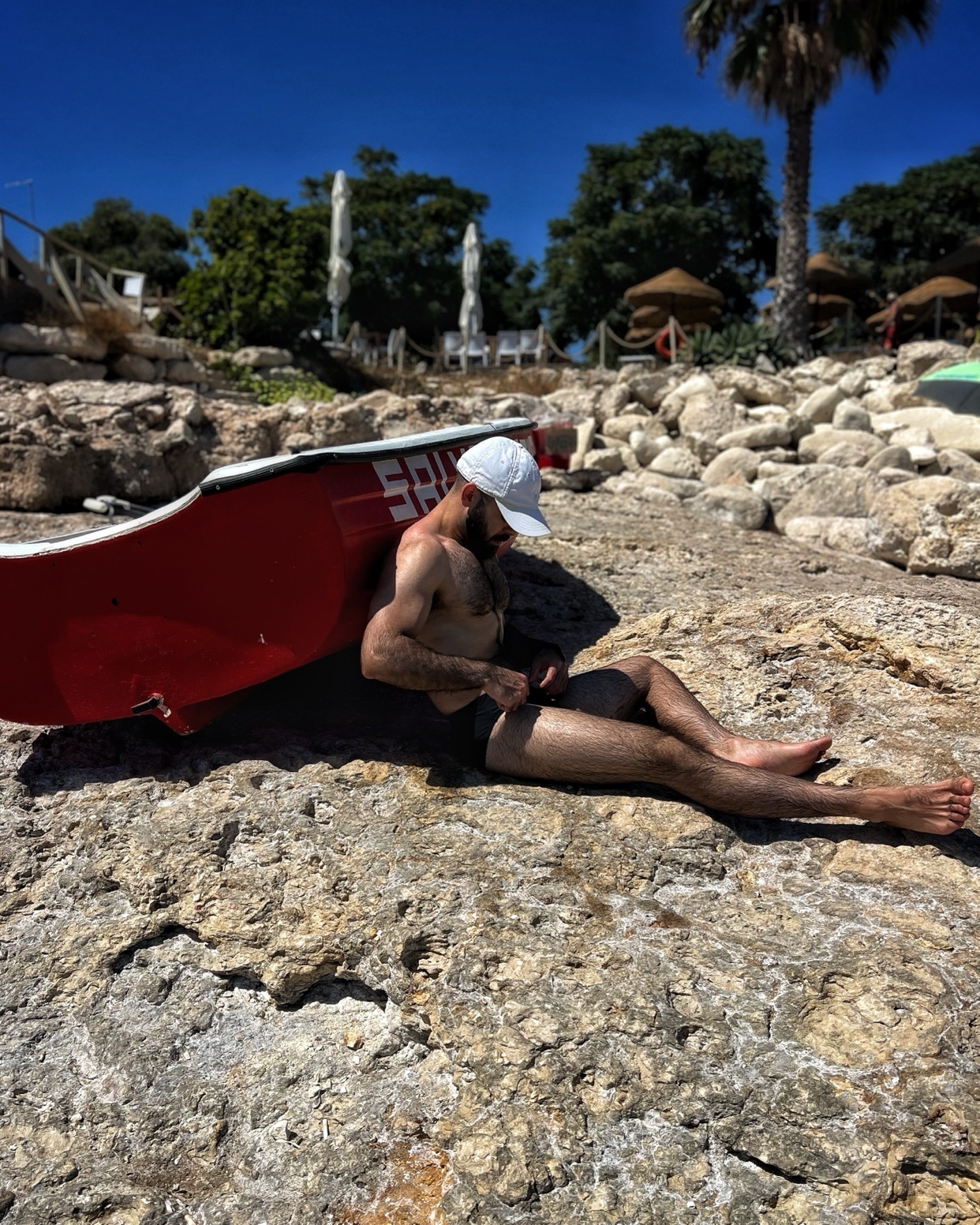 A man is shirtless, wearing a cap and swimwear, relaxing on a rocky beach. The photo is taken outdoors on a sunny day.