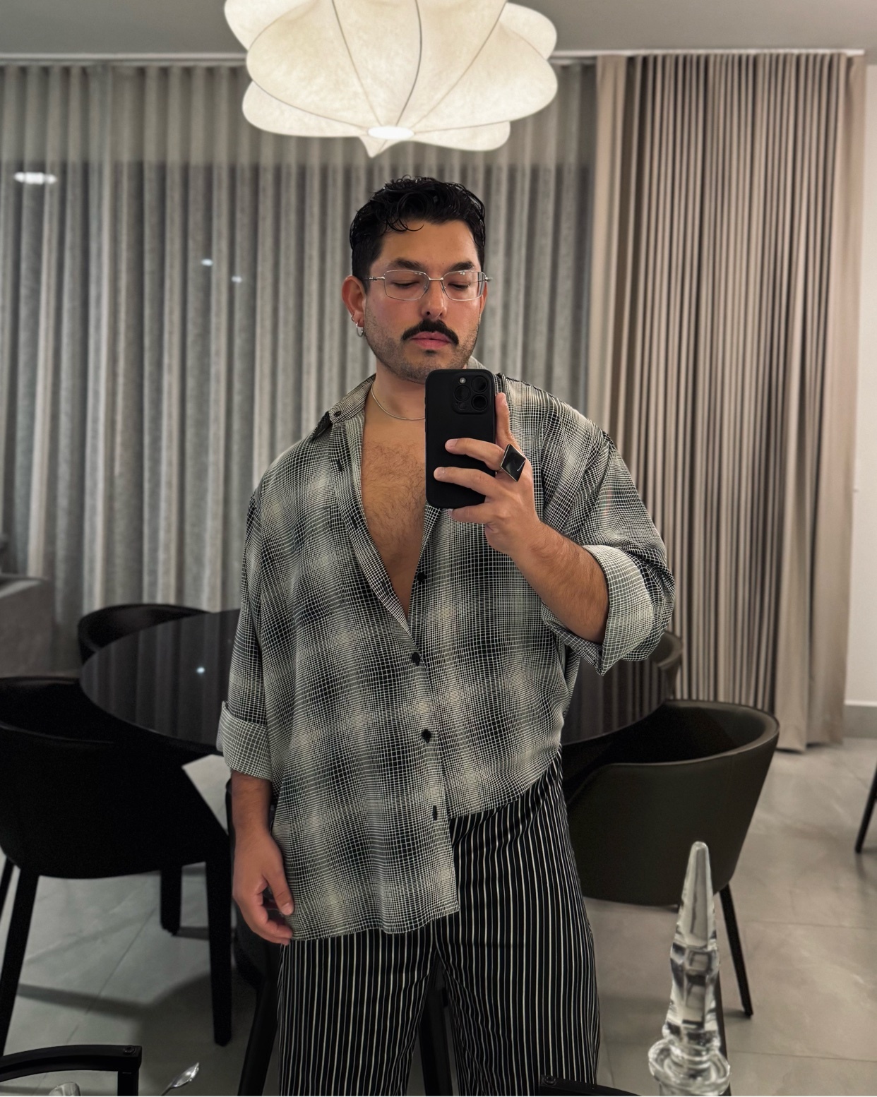 A man takes a mirror selfie, shirtless under an open plaid shirt. The photo is taken indoors, with modern decor in the background.