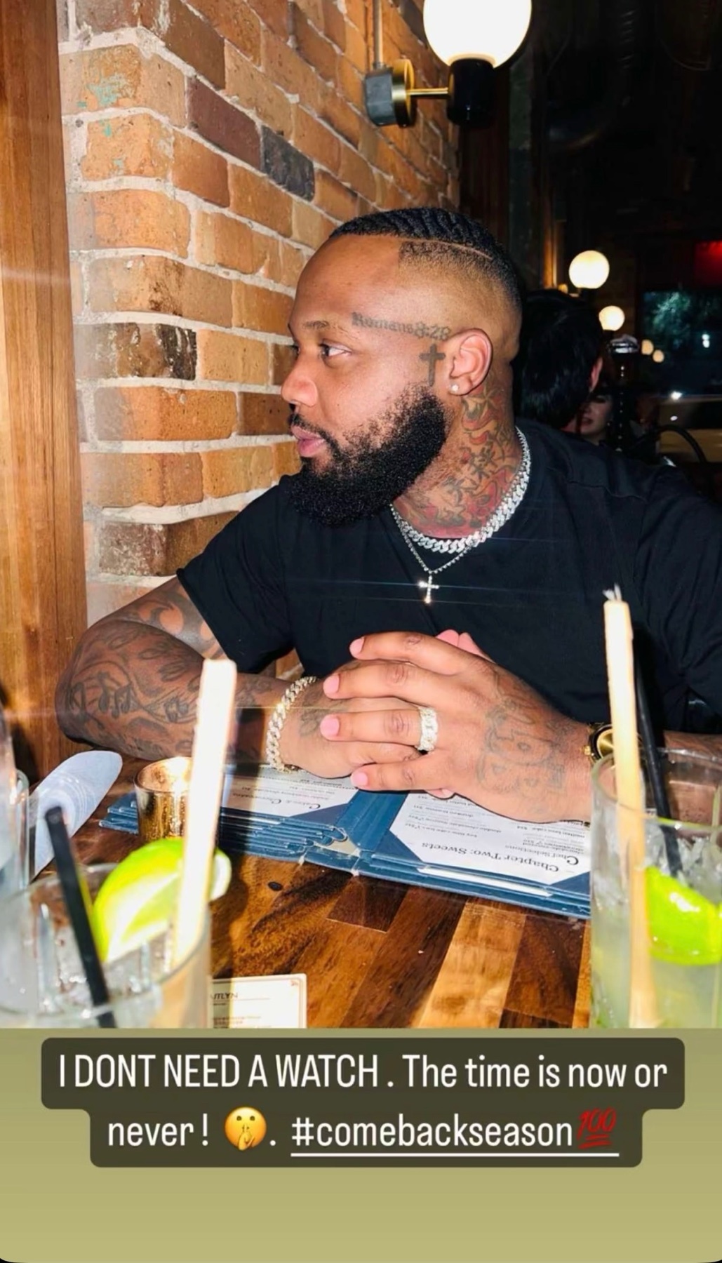 A man with a beard and braids sits at a table in a restaurant, looking to the side. He is wearing a black t-shirt and several pieces of jewelry, including a cross necklace and rings.