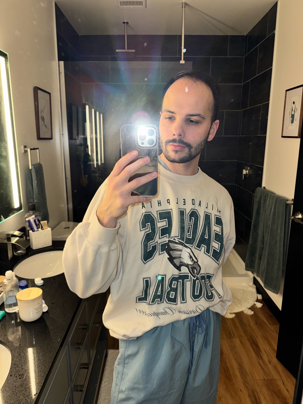 A man takes a mirror selfie in a modern bathroom. He is wearing an Eagles sweatshirt.