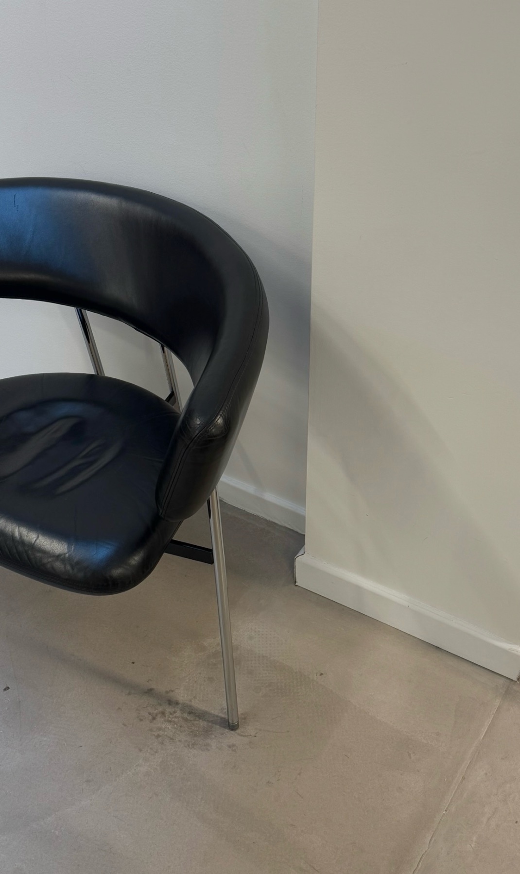 A black leather chair with metal legs sits on a tiled floor next to a white wall. There is a visible imprint on the seat of the chair.