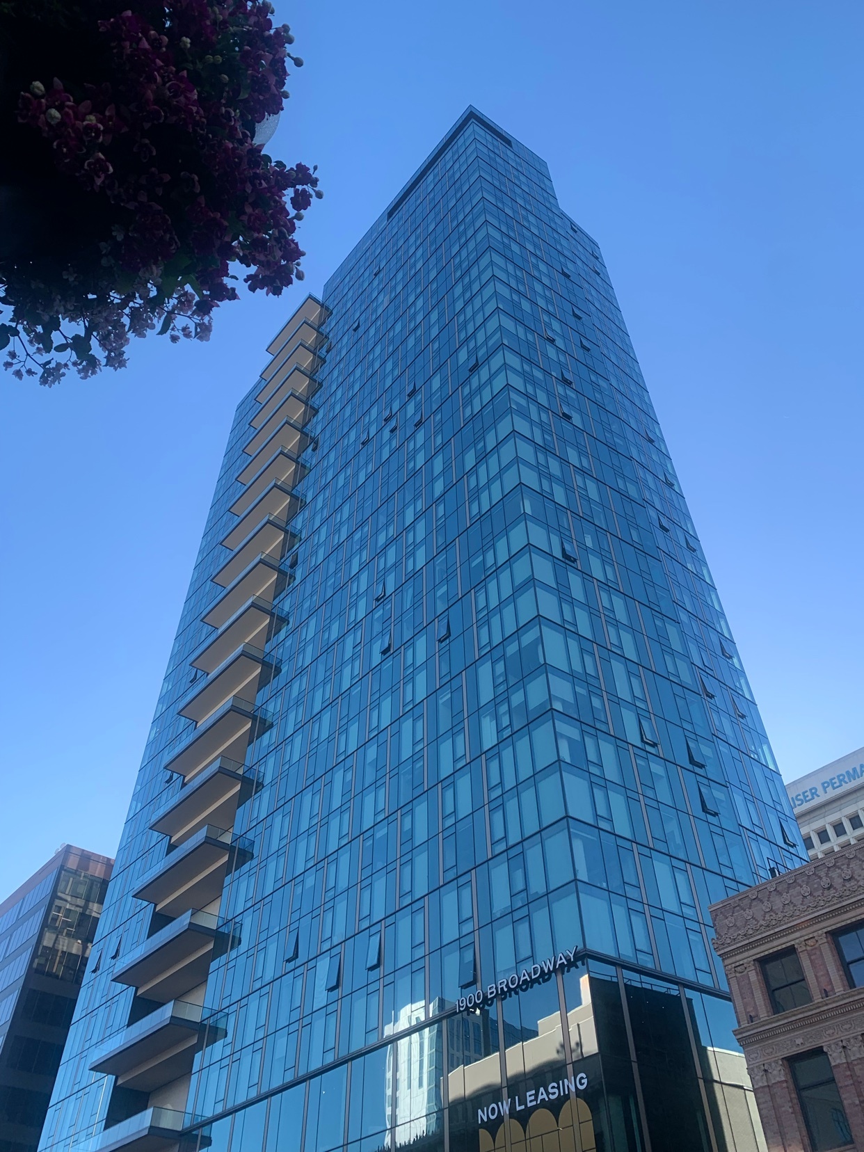 A modern glass skyscraper with balconies reaches towards a clear blue sky. The building features a 'Now Leasing' sign on its lower levels, indicating available apartments or office spaces.