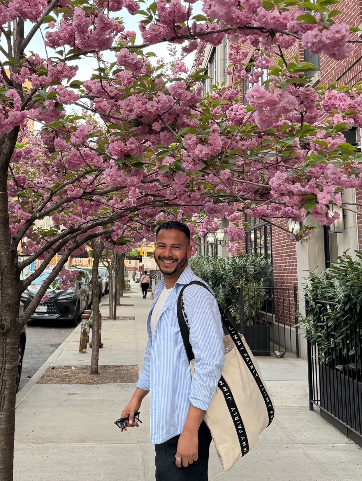 A man smiles while standing on a sidewalk under a blooming cherry blossom tree. He is wearing a light blue button-down shirt over a white t-shirt and dark pants, carrying a large tote bag over his shoulder.