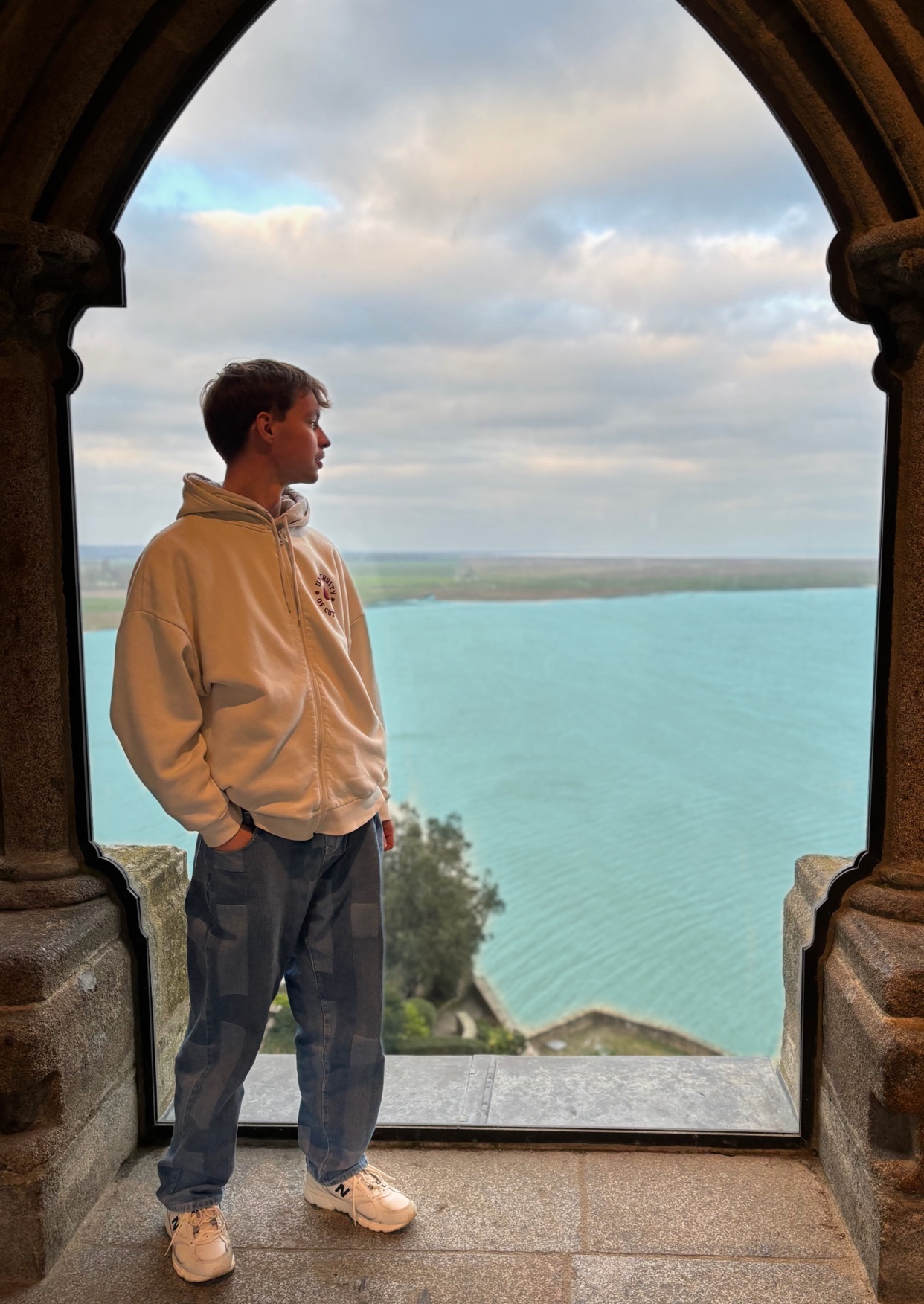 A man stands in a stone window, looking out at a scenic landscape. The photo captures a moment of contemplation with a beautiful view.