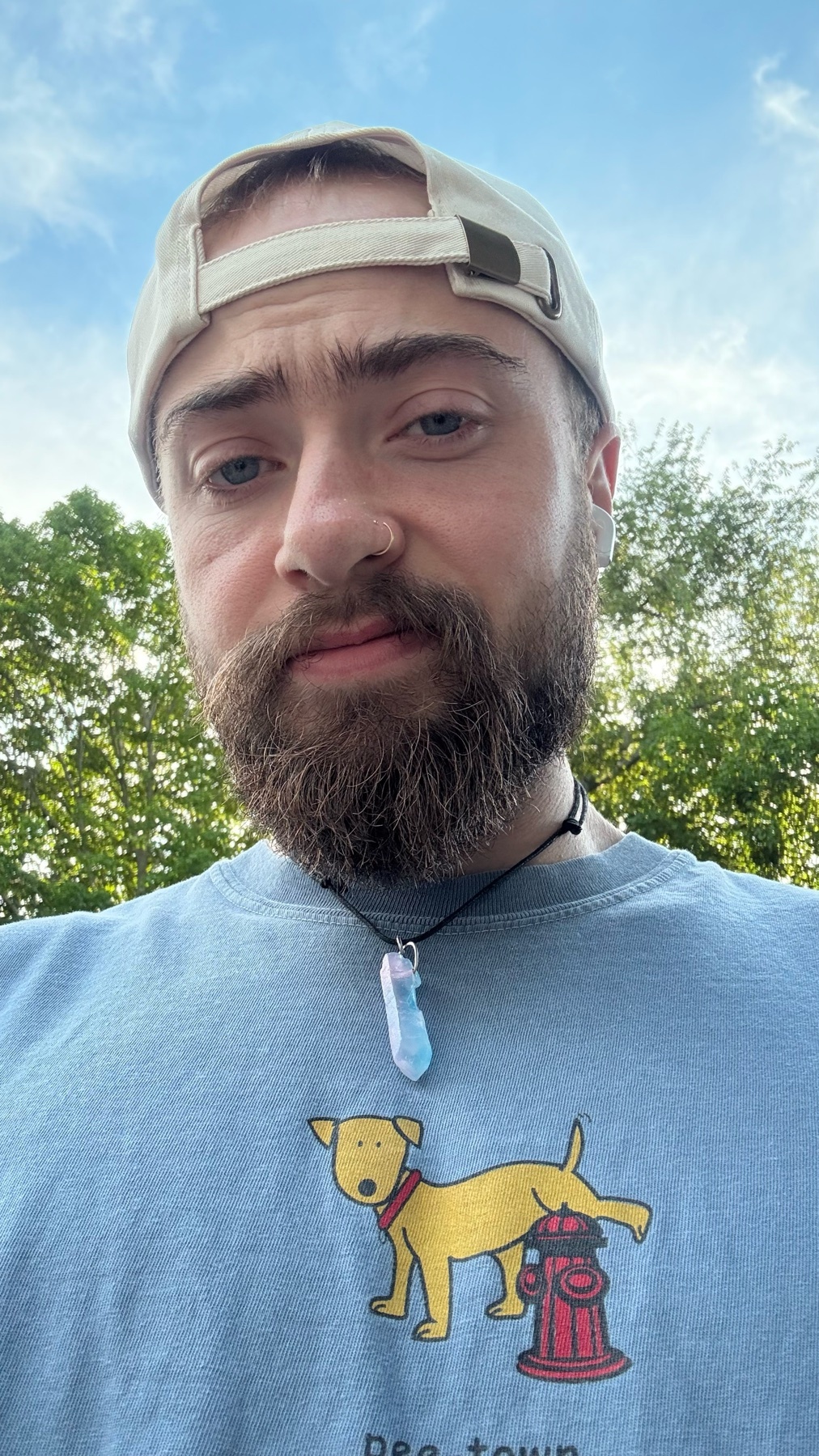 A man with a beard and a baseball cap takes a selfie outdoors. He is wearing a light blue t-shirt with a graphic of a dog peeing on a fire hydrant.