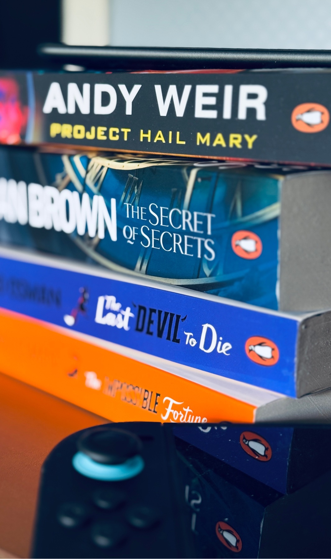 A stack of books is shown, with titles like 'Project Hail Mary' by Andy Weir and 'The Secret of Secrets' by Dan Brown visible. A video game controller rests in the foreground.