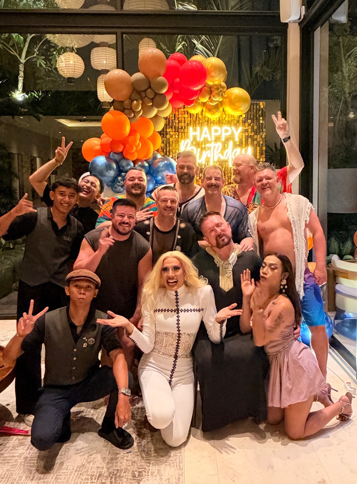 A group of people, including a drag queen, pose for a photo in front of a "Happy Birthday" balloon display. The group appears to be celebrating a birthday party.