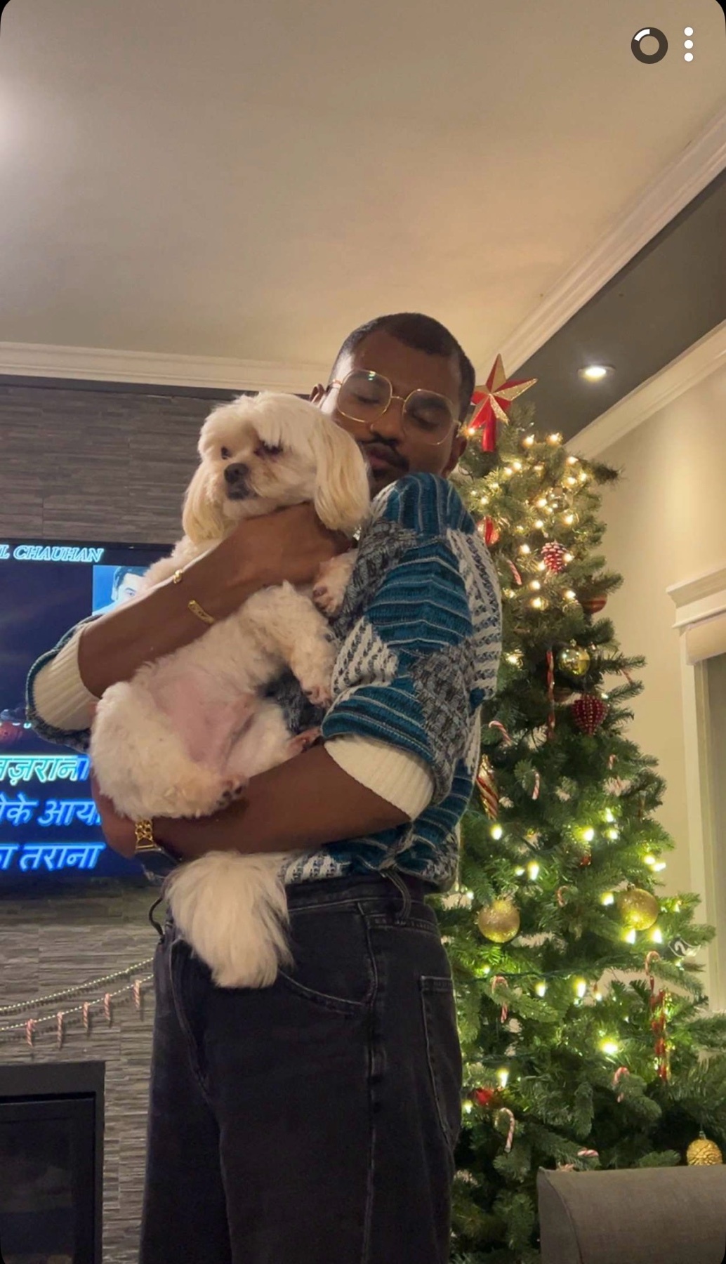 A man holds a small white dog in front of a Christmas tree. The man is wearing glasses and a sweater.