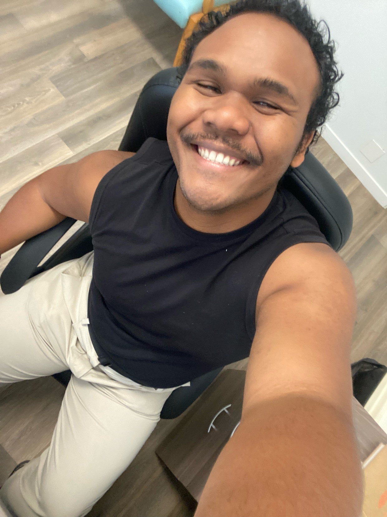 A man with curly hair and a mustache smiles at the camera in a selfie taken from above. He is wearing a black tank top and tan pants, sitting in a black office chair.