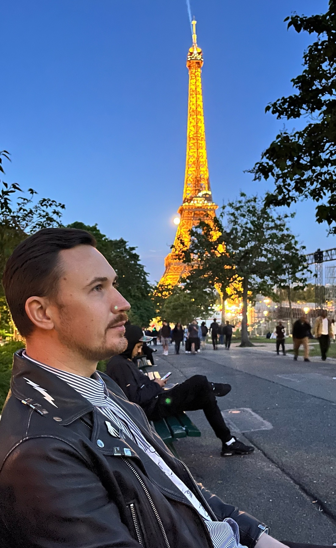 A man poses in front of the Eiffel Tower at night. He is wearing a leather jacket and a striped shirt.