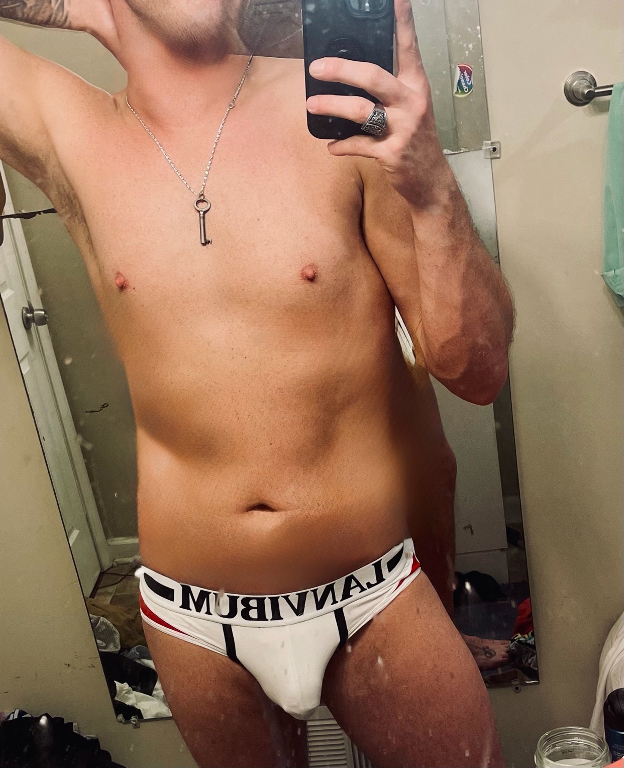 A man takes a mirror selfie in a bathroom, wearing only underwear. He has tattoos on his arm and is wearing a key-shaped necklace and a ring.