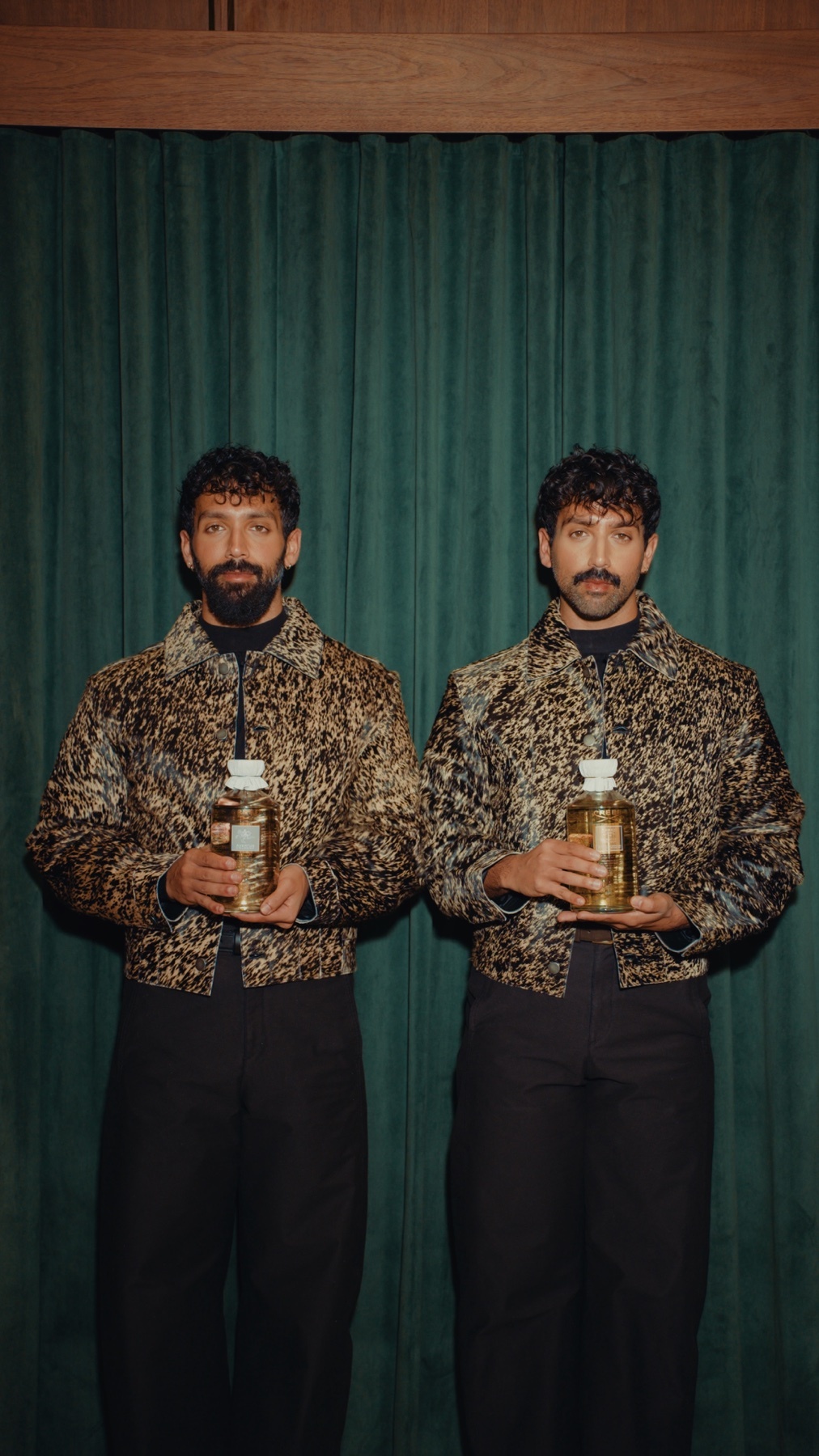 Two men pose with perfume bottles, wearing matching leopard-print jackets and black pants. The photo is a stylish portrait.