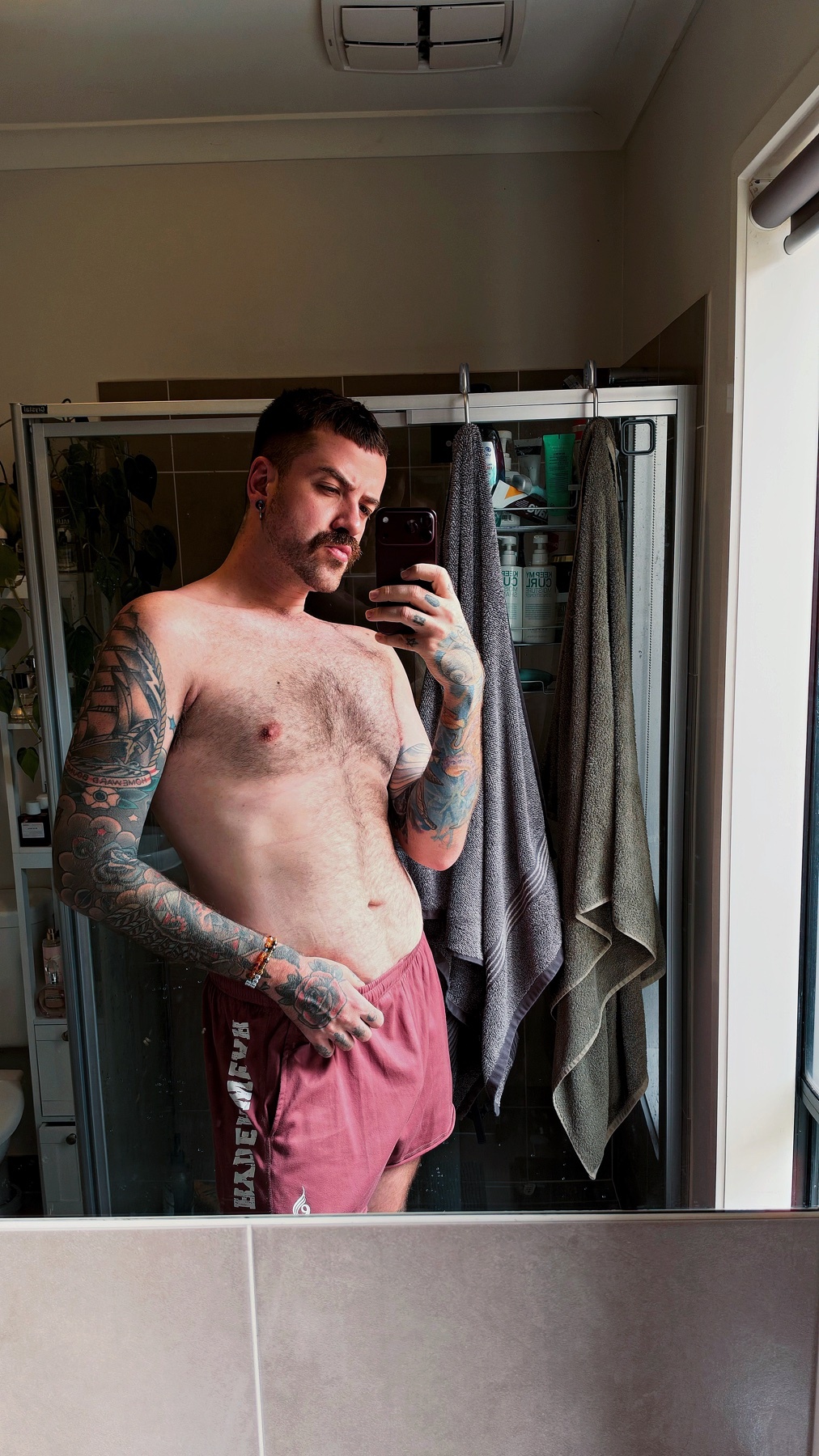 A tattooed man with a mustache takes a mirror selfie in a bathroom, wearing short shorts and no shirt.