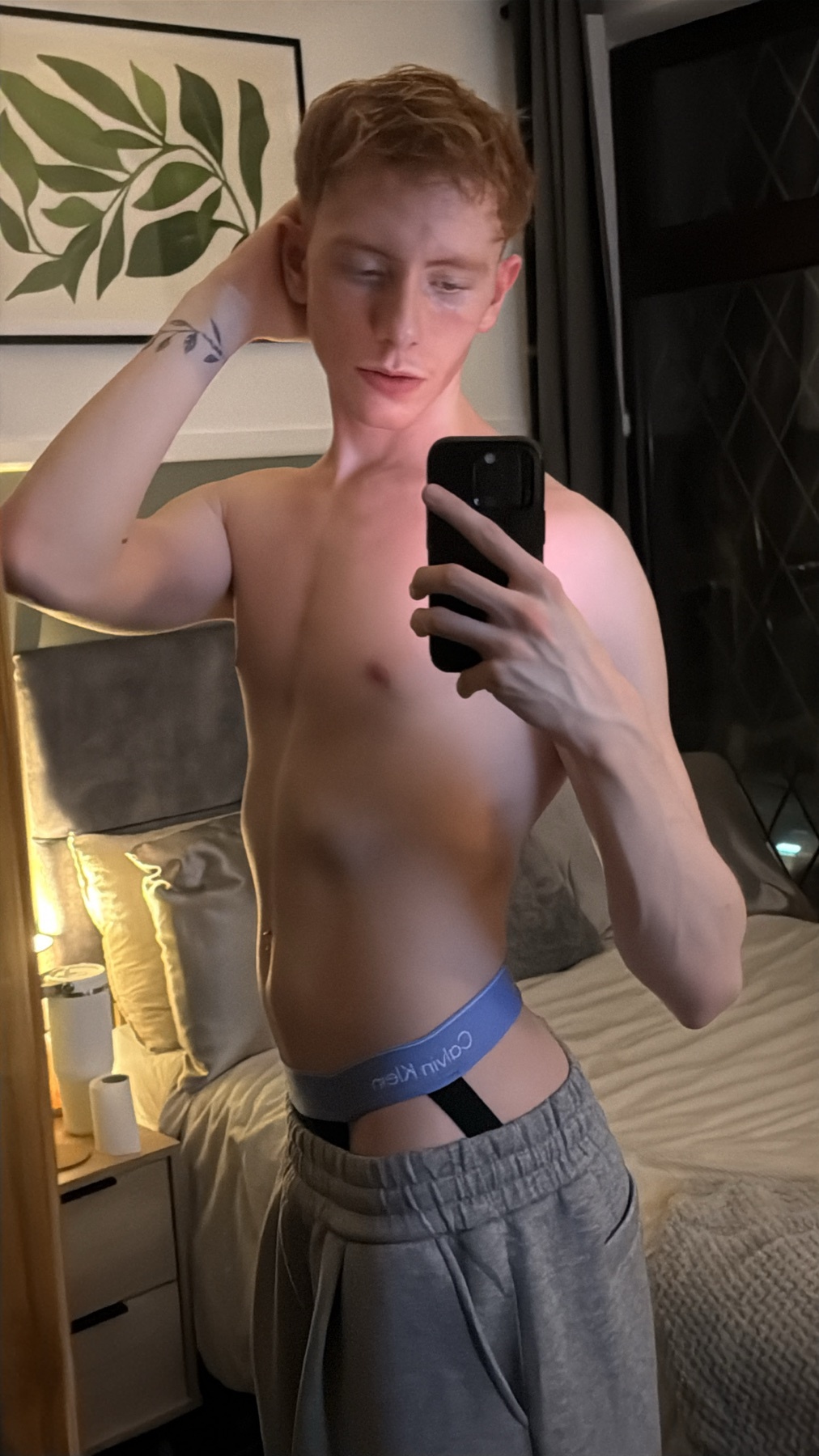A shirtless man takes a mirror selfie in his bedroom. He is wearing gray sweatpants and a blue Calvin Klein thong.
