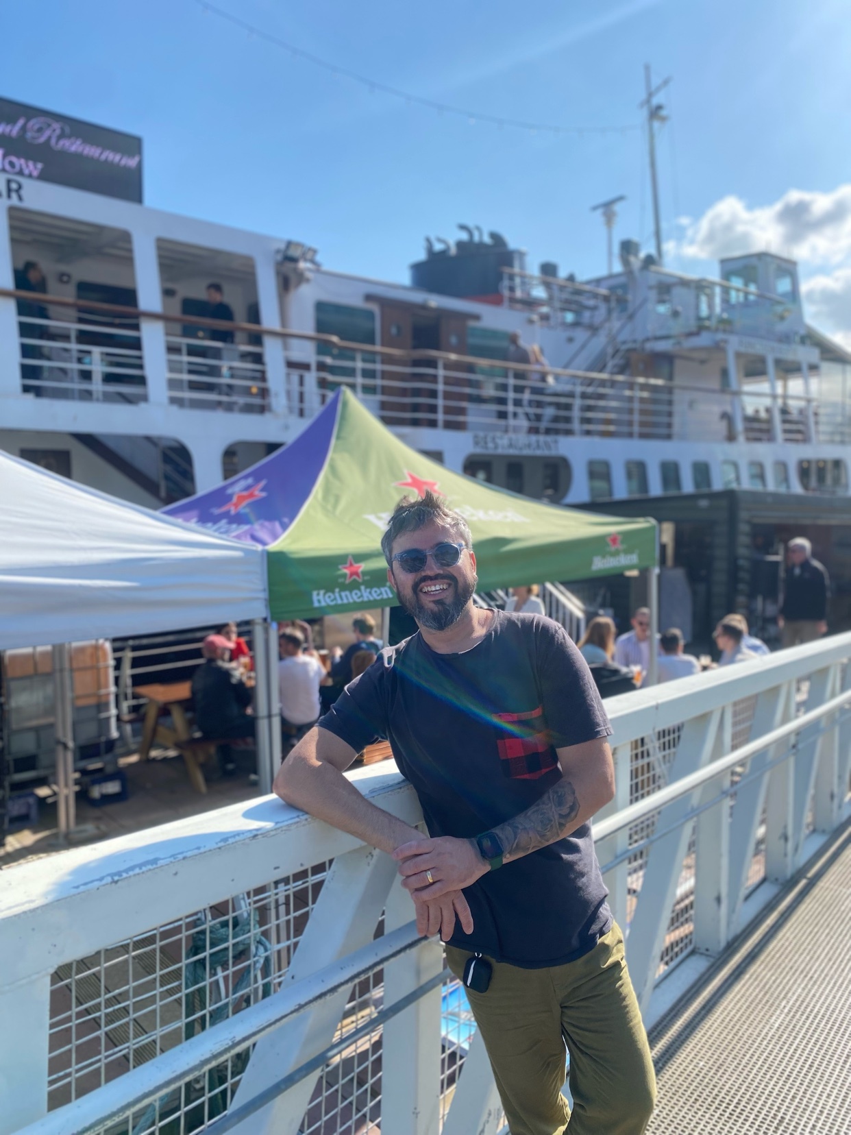 A man with a beard and sunglasses smiles at the camera while leaning on a railing on a sunny day. Behind him, a large boat is docked, with tents and people visible.