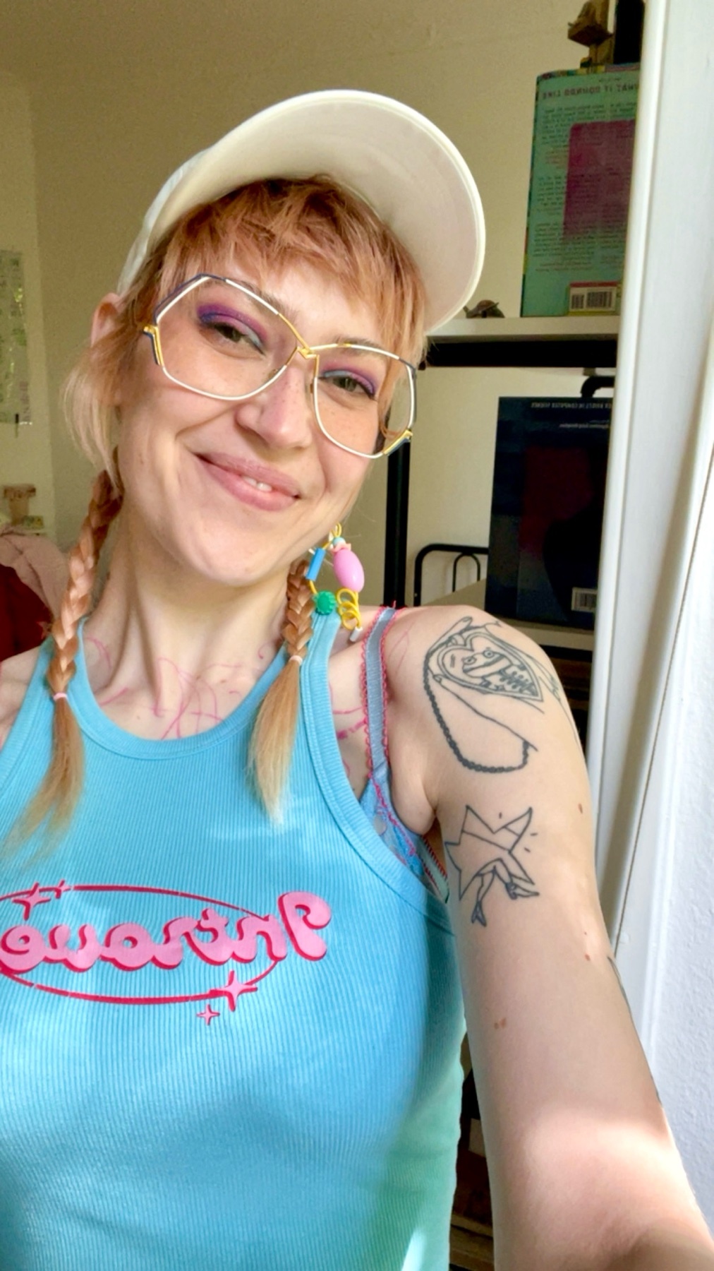 A woman with blonde hair in pigtails and large glasses smiles at the camera. She is wearing a blue tank top and a white baseball cap.