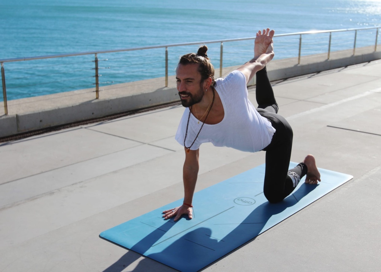 A man with a beard and man bun performs a yoga pose on a mat outdoors with the ocean in the background.