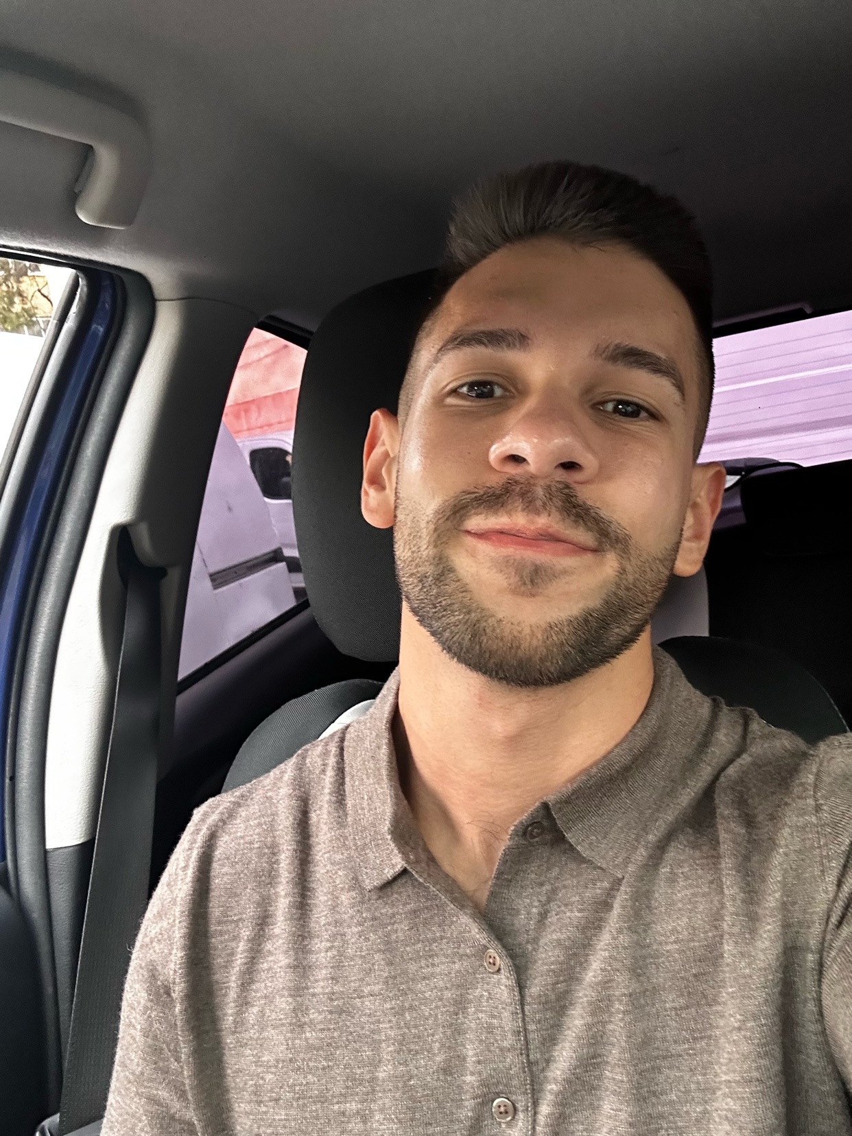 A man with short dark hair and stubble is looking at the camera while sitting in the driver's seat of a car.