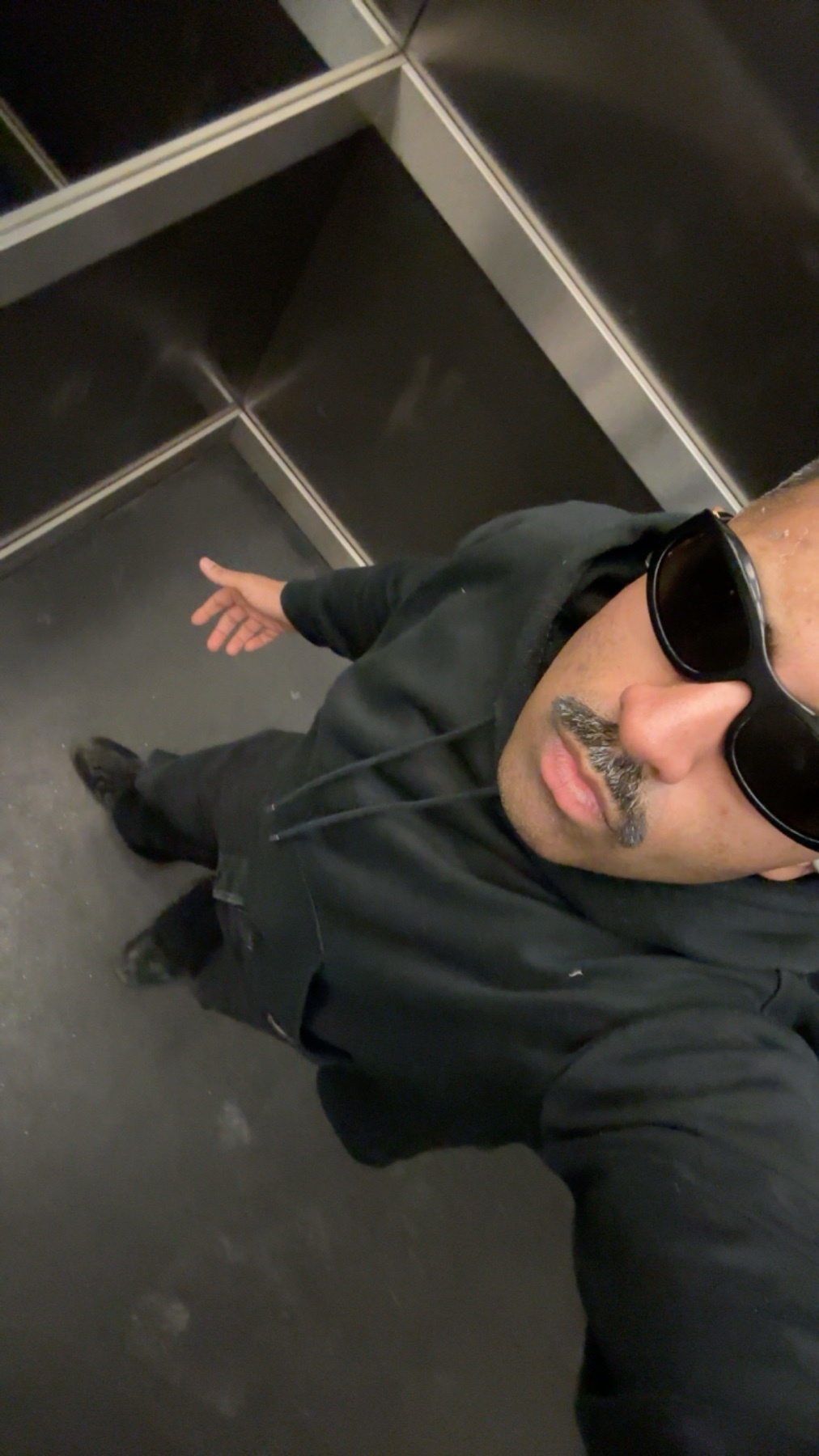A man in sunglasses and a hoodie takes a selfie in an elevator.