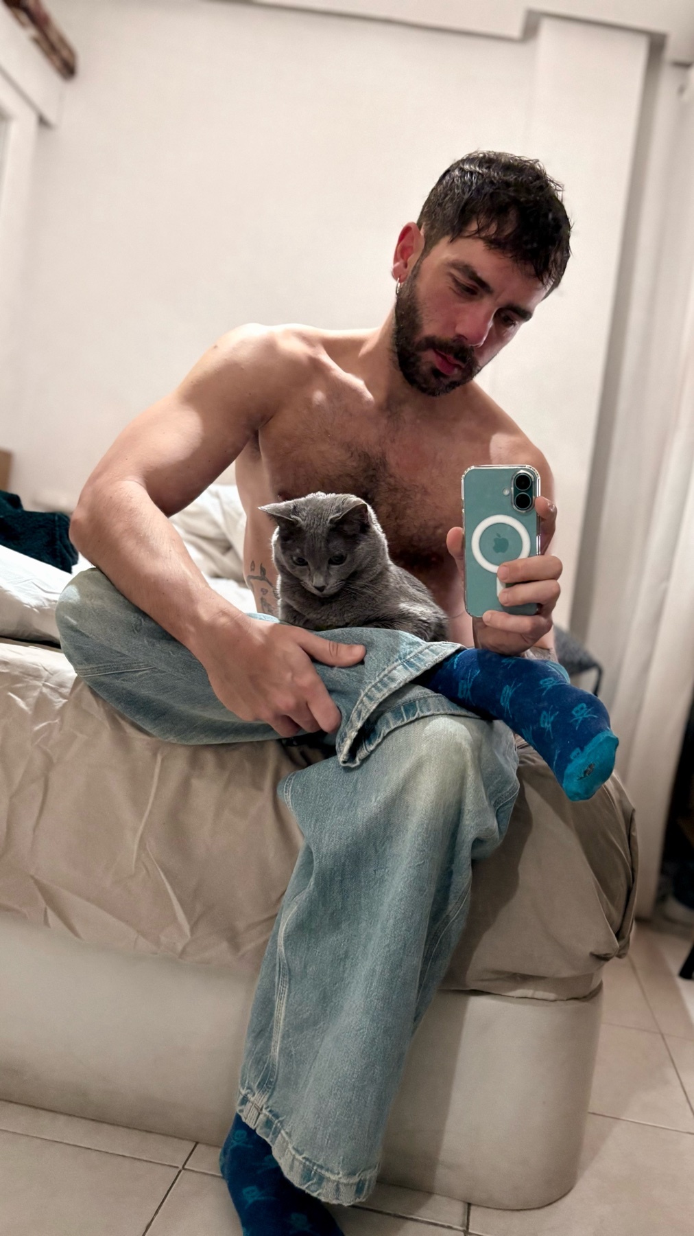 A man sits shirtless, holding a cat on his lap. He is taking a selfie, and has a blue cast on his leg.
