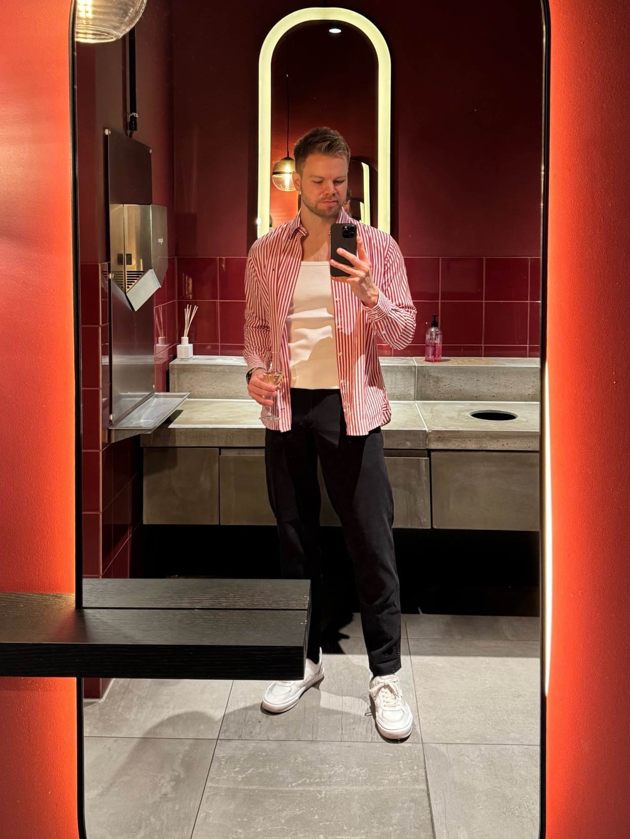 A man takes a mirror selfie in a stylish bathroom. He wears a red and white striped shirt, a white undershirt, black pants, and white sneakers.