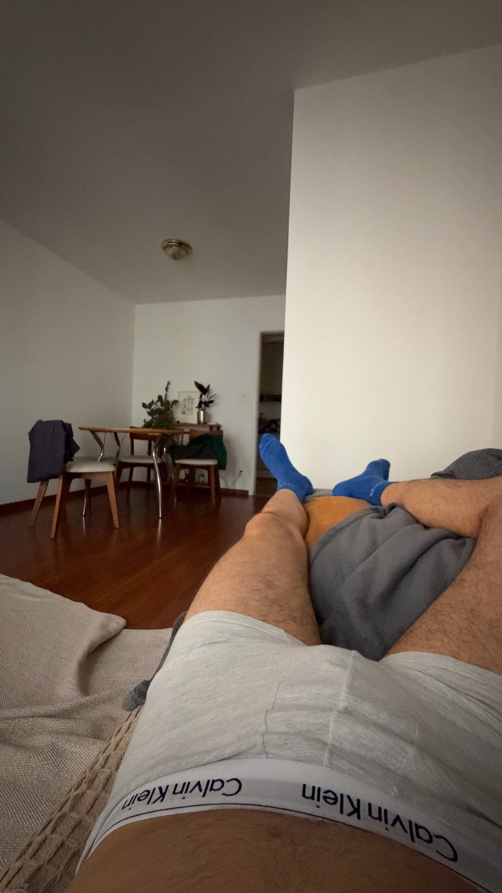 A close-up, low-angle shot of a man lying down in his underwear and socks. The shot is taken from his perspective, looking towards a dining table and chairs in the background.