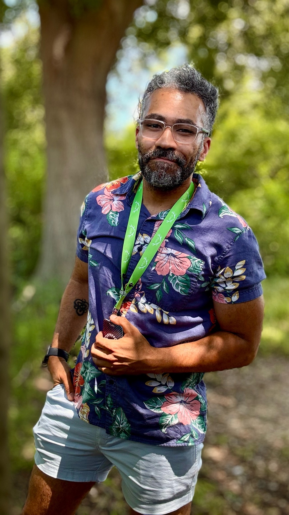 A man with glasses and a beard poses outdoors wearing a colorful Hawaiian shirt and light-colored shorts. He is standing in front of a blurred background of trees and greenery.