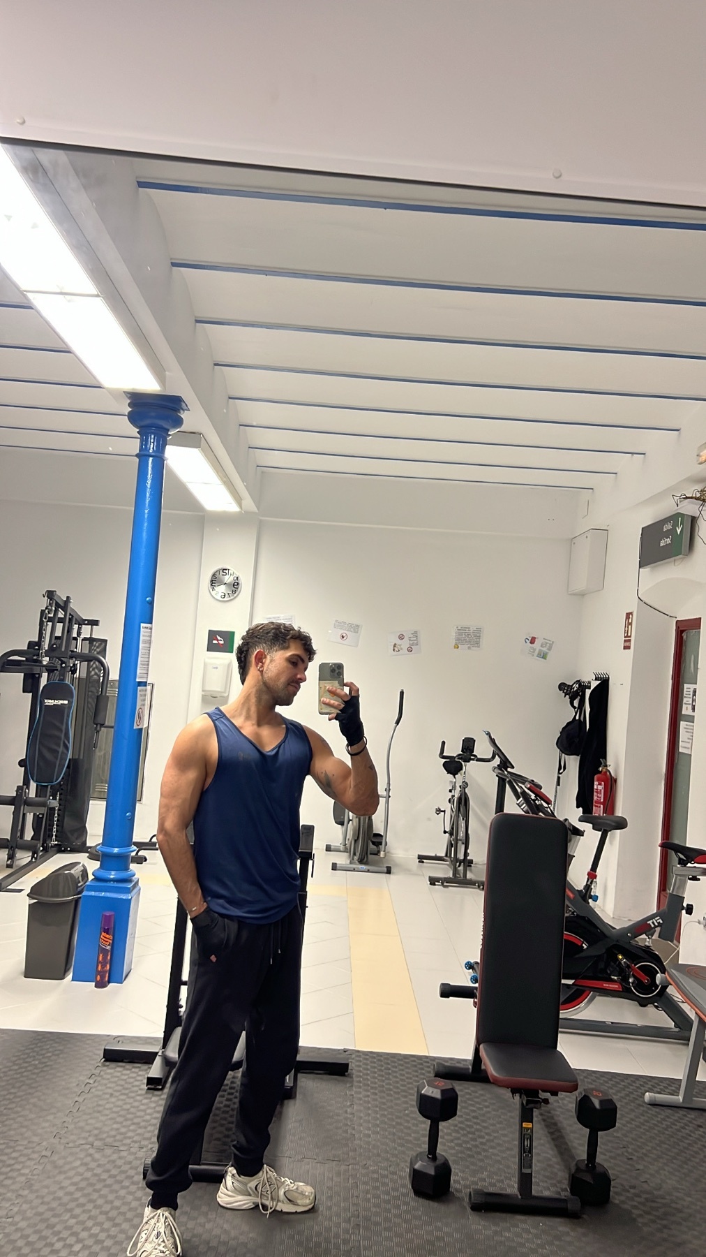 A man takes a mirror selfie in a gym, flexing his arm. He is wearing a blue tank top and black pants.