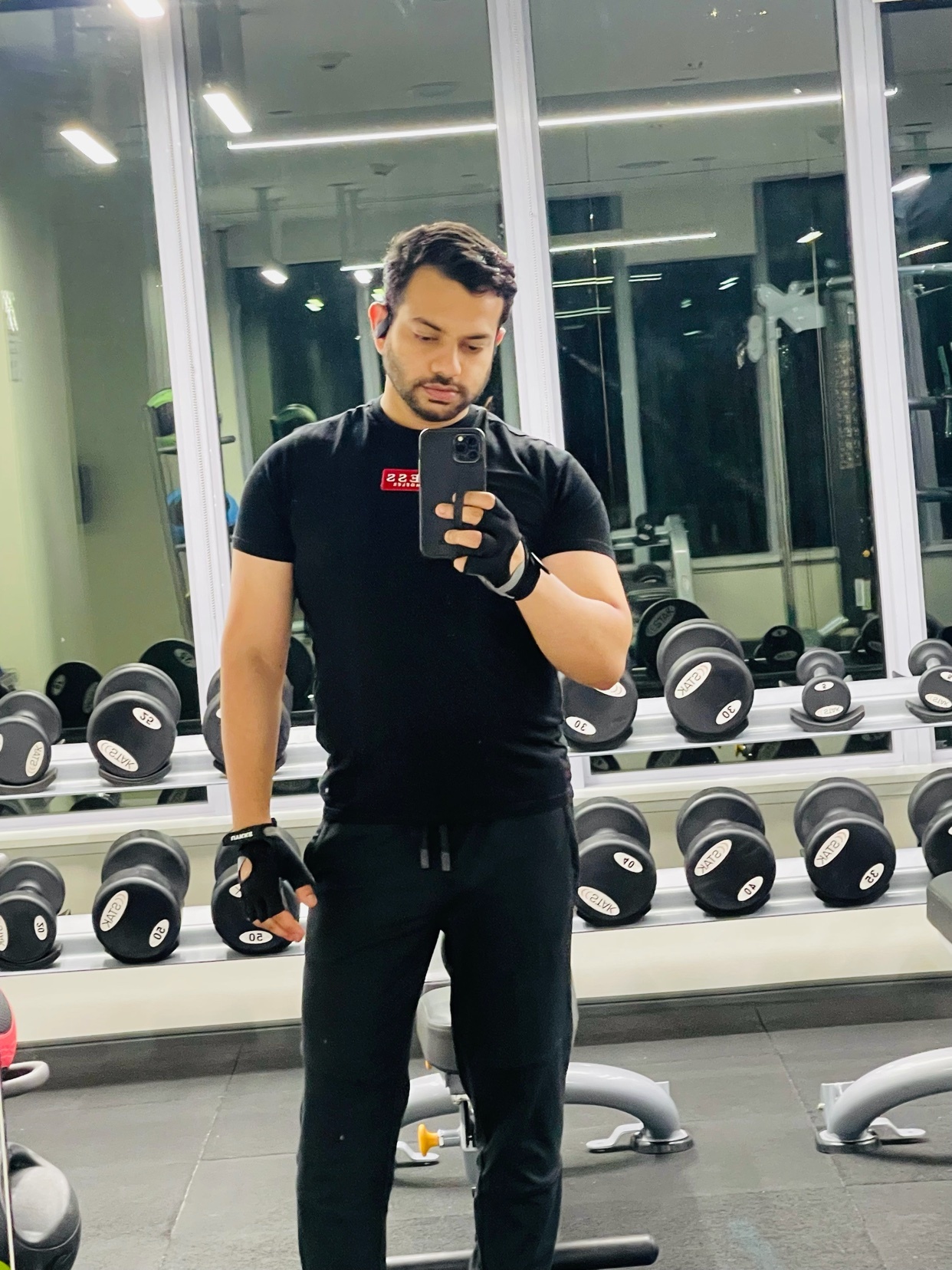 A man takes a mirror selfie in a gym, surrounded by weights. He's wearing a black t-shirt and gloves, ready for a workout.